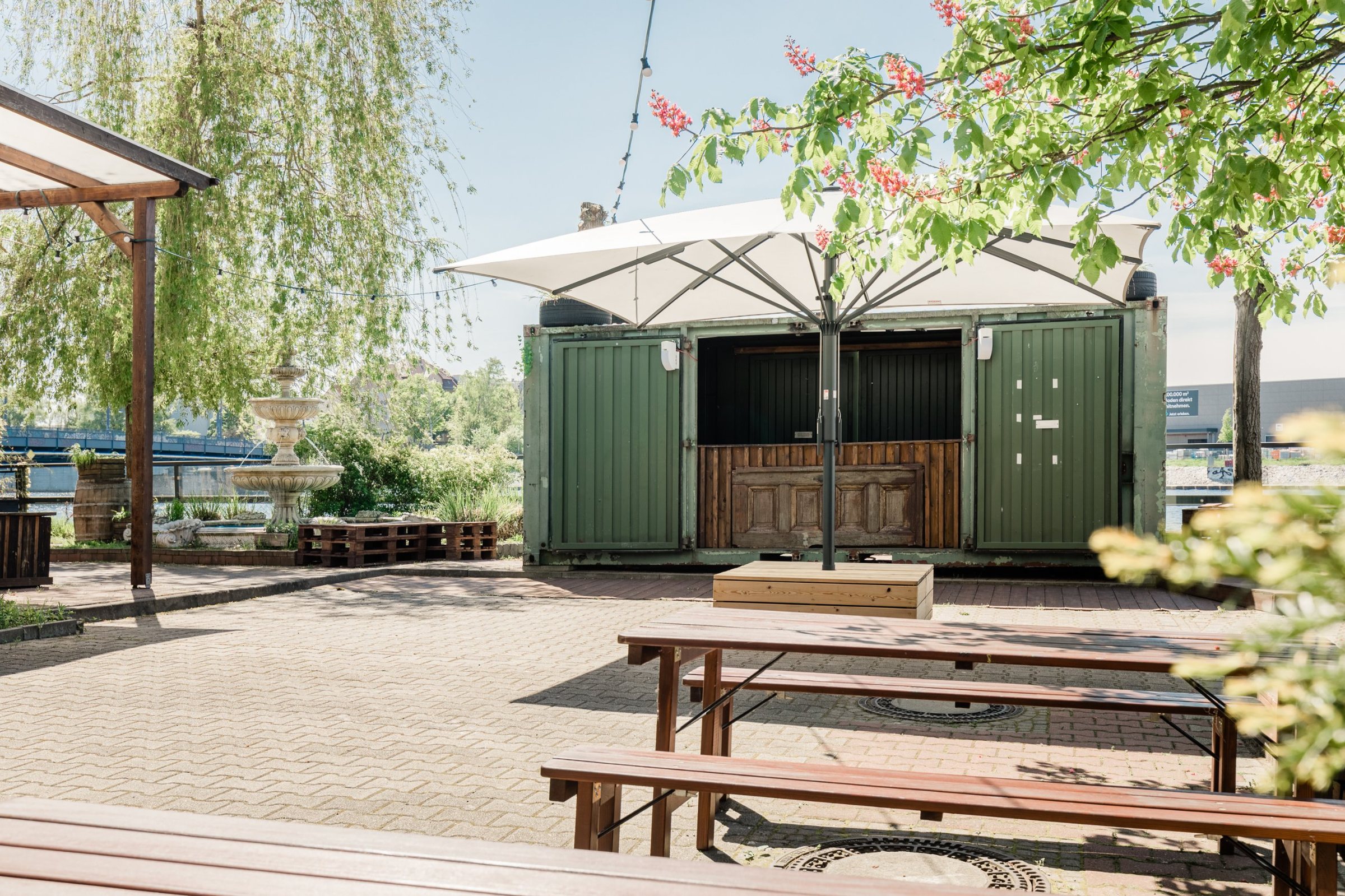 Innenhof mit Container-Bar, Sonnenschirm, Bierbänken und Brunnen