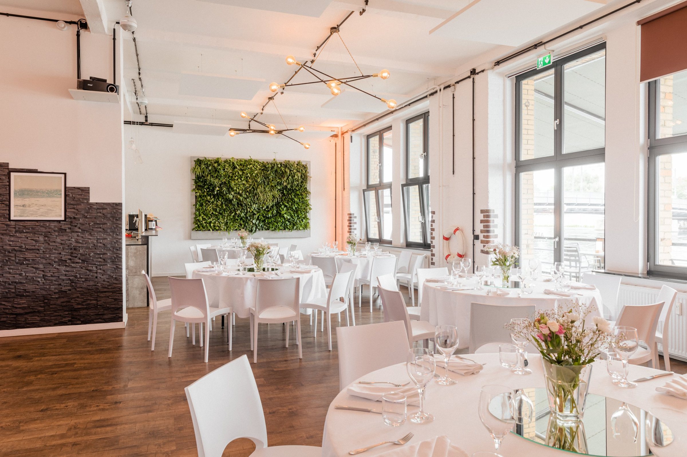ALVA Lounge mit runden, festlich gedeckten Tischen, Pflanzenwand und Fenstern zur Spree